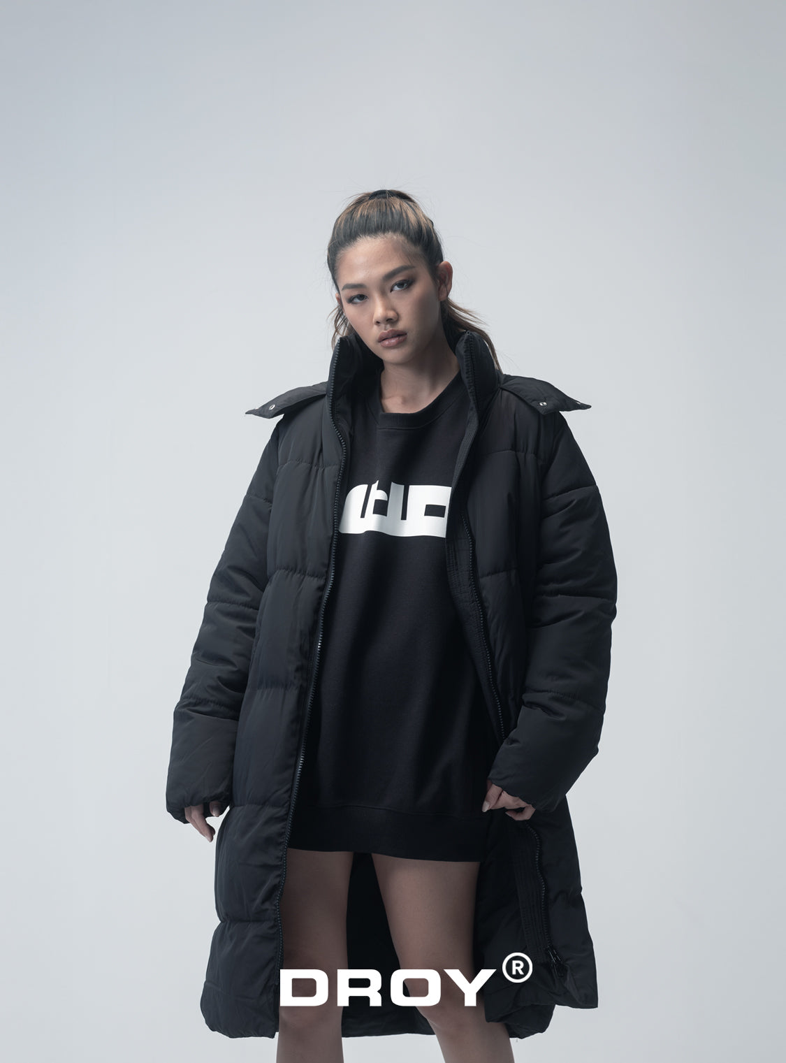 Person wearing a black puffer coat and black dress with a logo, standing against a white background.