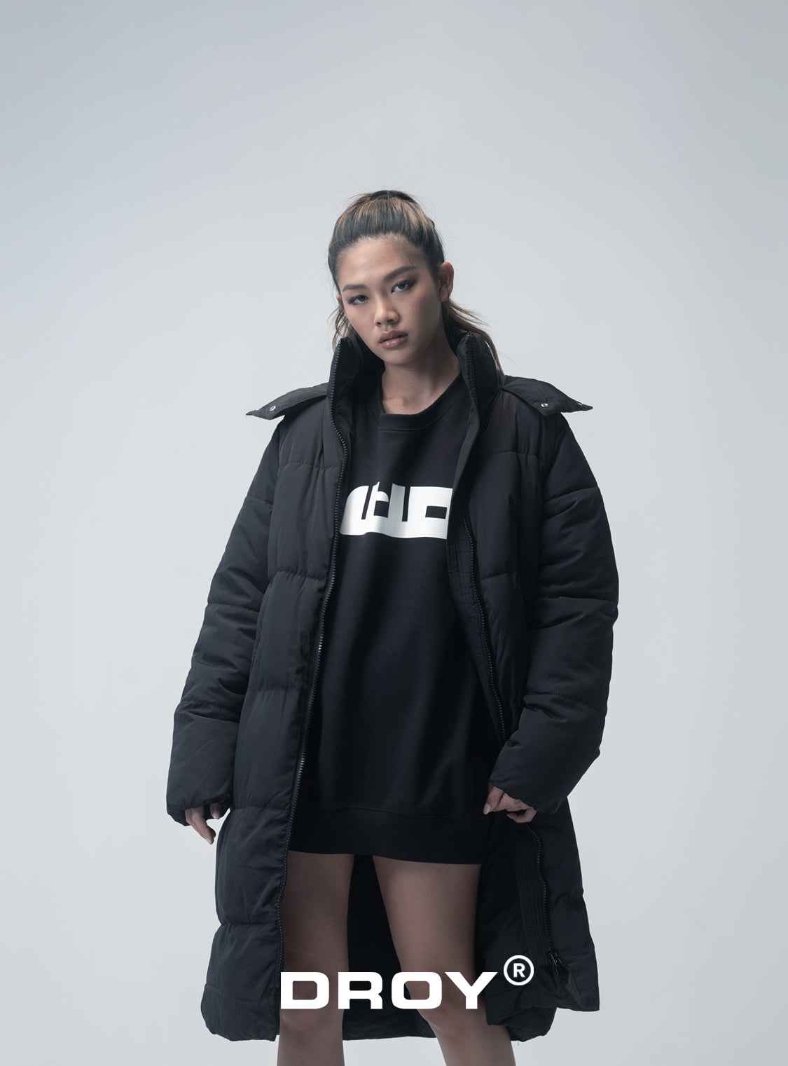 'DROY' Person wearing a black puffer coat and black dress with a logo, standing against a white background.