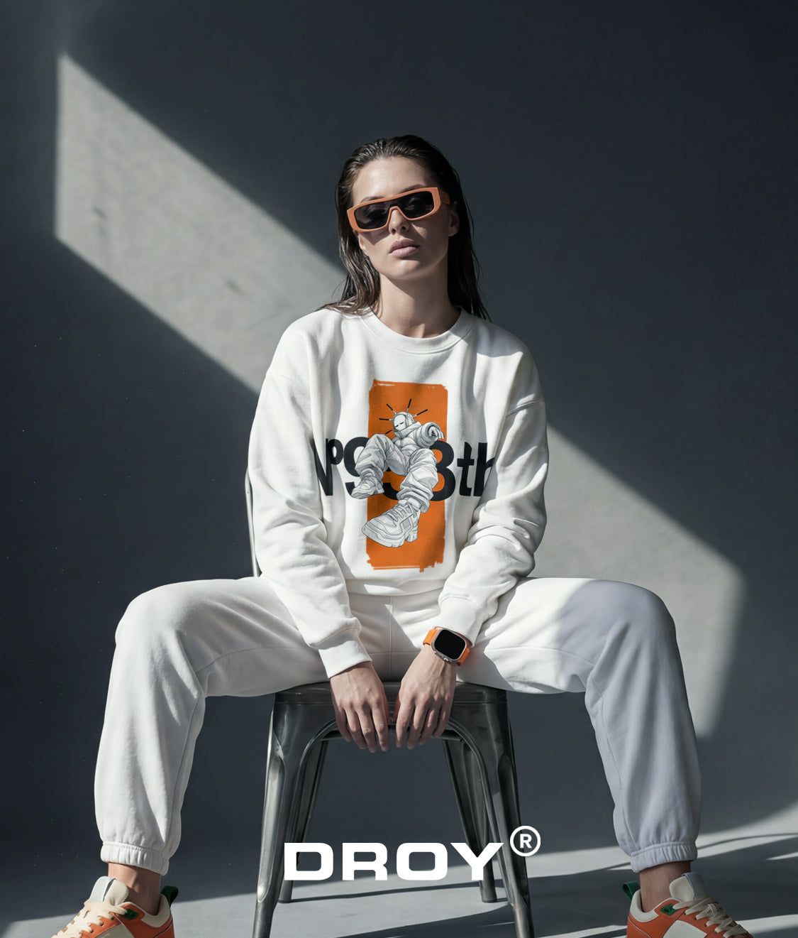 'DROY' Person wearing a white sweatshirt with a graphic design, sitting on a stool against a dark wall.
