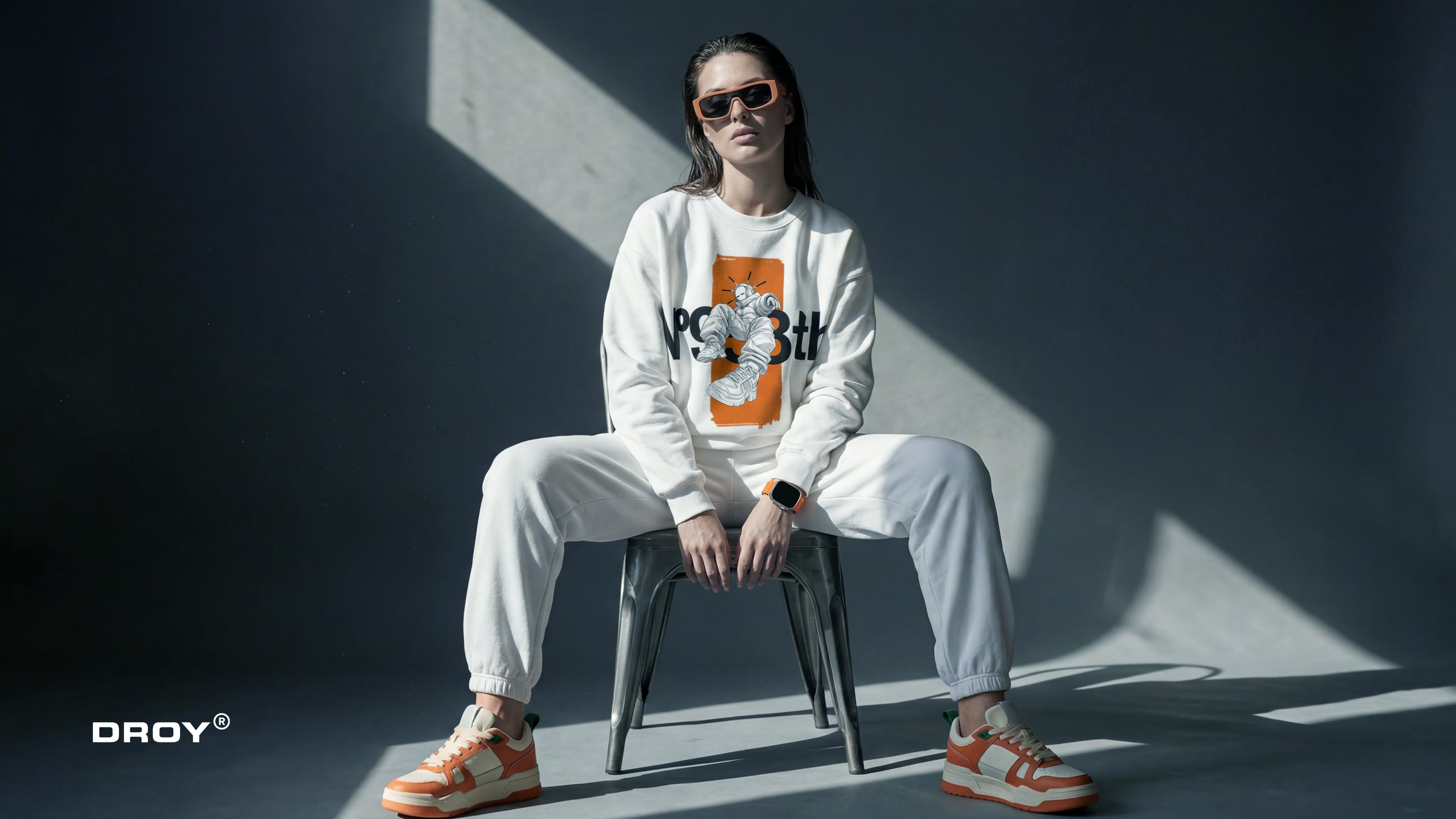 Person sitting on a chair wearing a white sweatshirt with a graphic design, sunglasses, and white pants. The brand 'DROY' is visible.
